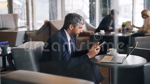 一个人坐在咖啡馆里用智能手机触摸屏幕的中年企业家视频的预览图