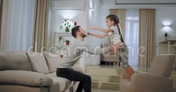 在一个宽敞的客厅里一个年轻的父亲和他的小女儿在沙发上玩耍小女孩跳过去视频的预览图