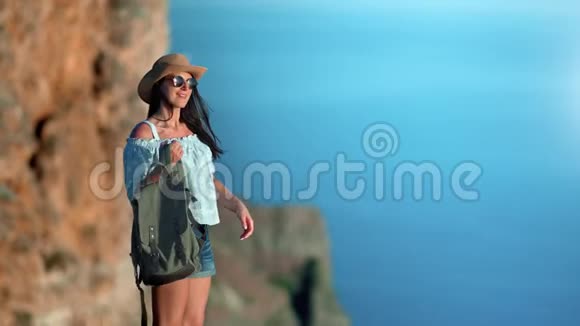 美丽的微笑放松的年轻女子戴着帽子戴着太阳镜背着背包欣赏着令人惊叹的大海视频的预览图
