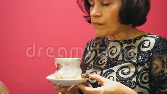 年长的黑发女人正在喝一杯茶吃红色背景的糖果视频的预览图