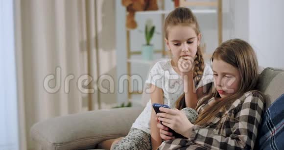 一幅可爱的两个姐妹在智能手机上玩耍的肖像集中在一个宽敞的客厅里坐在沙发上视频的预览图