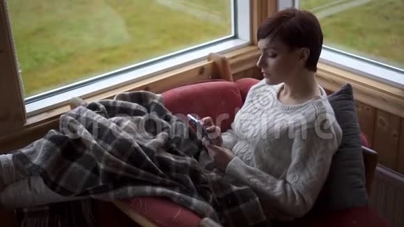 少妇在雨天坐在家里的椅子上使用智能手机视频的预览图