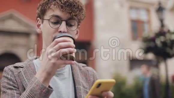 在坐在长凳上的时候用智能手机和喝咖啡把戴眼镜和夹克的年轻潮人关上视频的预览图
