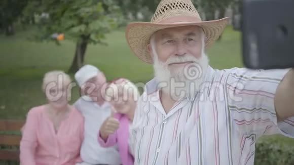 戴着帽子留着灰色胡子的成年男子在公园里坐在长凳上的朋友的背景下自拍视频的预览图