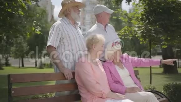 两对可爱的成熟夫妇在夏天公园的长凳上聊天和微笑双人约会的高级夫妇友好视频的预览图