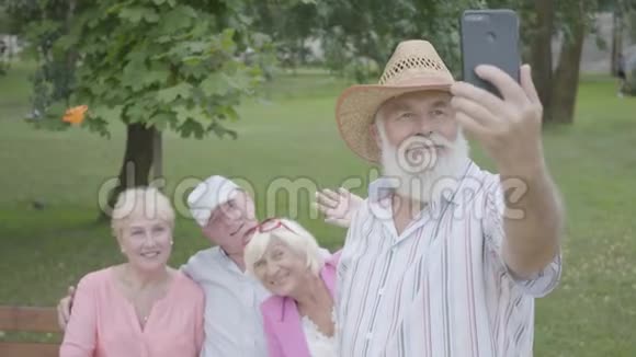 戴着帽子和灰色胡子的老人在公园里坐在长凳上的朋友的背景下自拍二视频的预览图