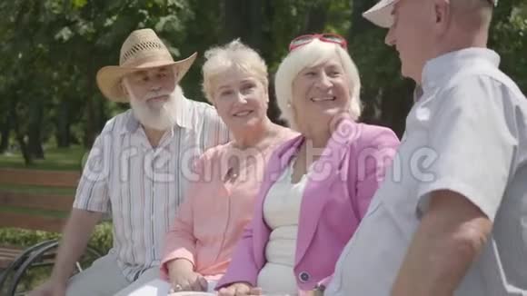 两对可爱的成熟夫妇在夏天公园里坐在长凳上聊天和微笑老年夫妇双人约会视频的预览图
