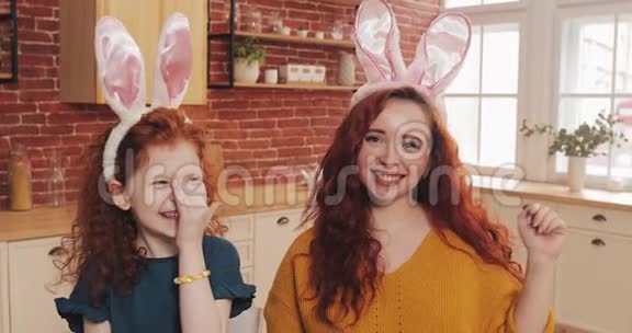 复活节快乐性格开朗的红发小女孩和妈妈一起穿着兔耳制作视频聊天或与亲戚聊天视频的预览图