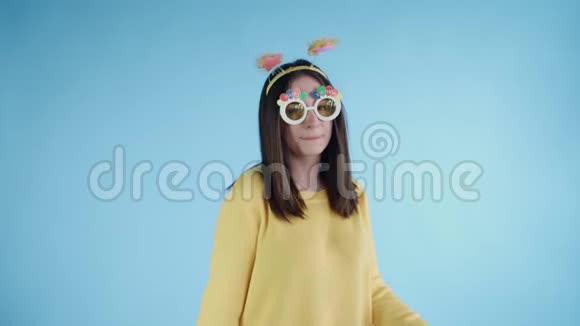 漂亮的年轻女子戴着滑稽的帽子和眼镜独自在蓝色的背景上跳舞视频的预览图