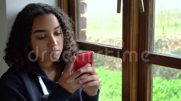 非洲裔美国少女沮丧地坐在窗户旁用手机或智能手机视频的预览图