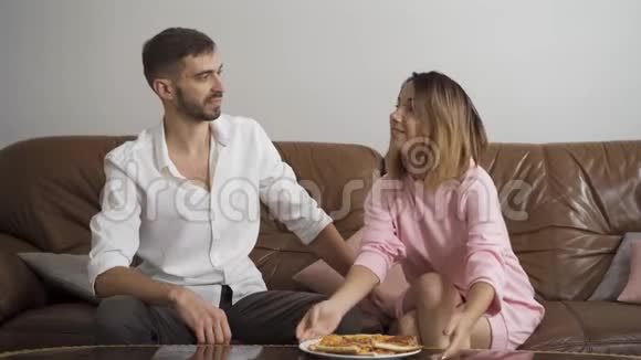 穿着便服的女人用美味的自制披萨喂她丈夫坐在皮革沙发上新婚夫妇快乐视频的预览图