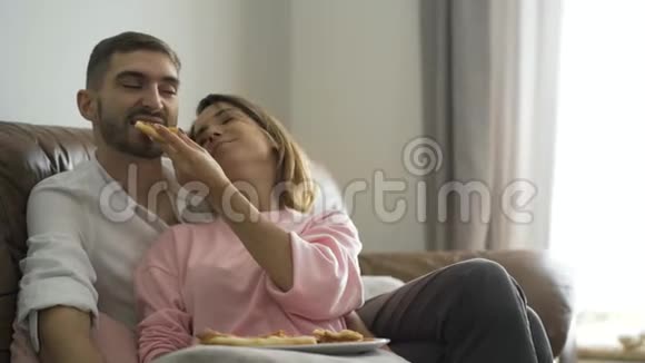 穿着便服的女人用美味的自制披萨喂她丈夫坐在皮革沙发上新婚夫妇快乐视频的预览图
