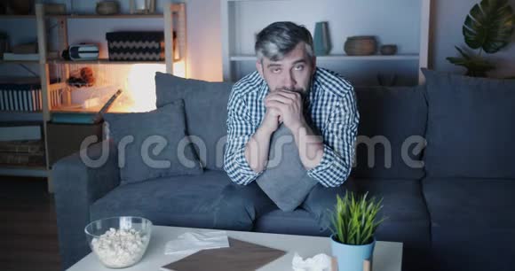 一幅悲伤的男人在晚上在家看电视时用纸巾视频的预览图