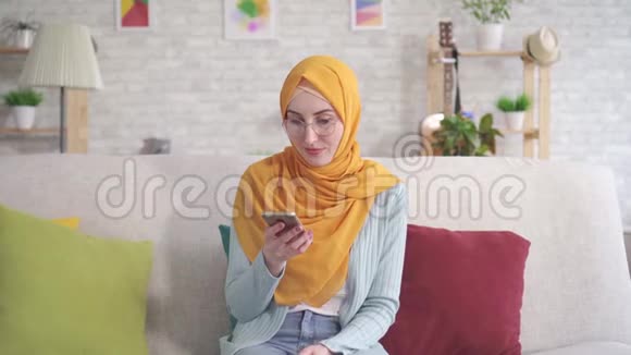戴头巾的年轻穆斯林妇女正微笑着拿着智能手机和银行卡坐在客厅的沙发上视频的预览图