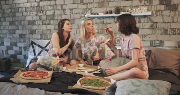漂亮的年轻女士穿着睡衣在家吃披萨互相聊天坐在床上微笑着视频的预览图
