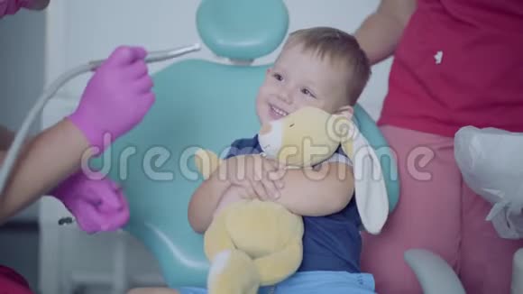 牙医戴着医用口罩和手套准备检查坐在椅子上的无忧无虑的小男孩的牙齿淘气鬼视频的预览图