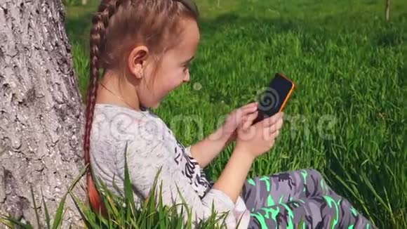 一个可爱的小女孩坐在树下的草地上手里拿着电话视频的预览图