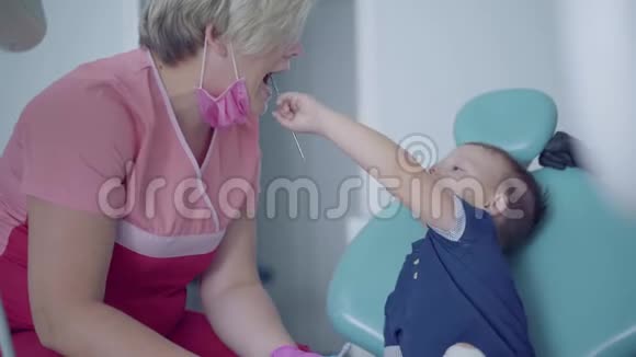 牙医戴着医用口罩和手套准备检查坐在椅子上的无忧无虑的小男孩的牙齿淘气鬼视频的预览图