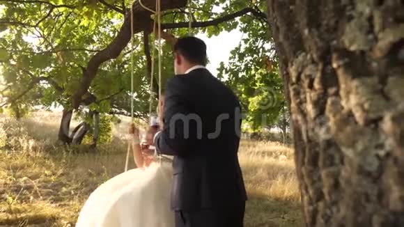 可爱的夫妇在公园里骑着秋千在树枝上的绳子上摆动愉快的年轻家庭蜜月旅行视频的预览图