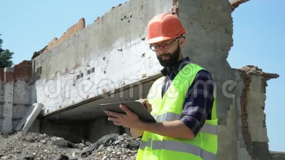 戴着头盔和反光背心的长胡子建筑工人在被摧毁的建筑背景上使用平板电脑为准备视频的预览图