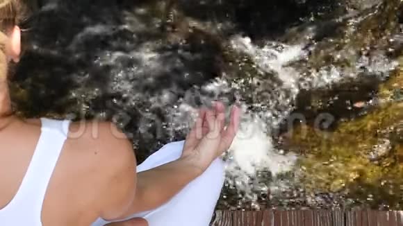 年轻女子练习瑜伽和冥想慢动作坐在瀑布的莲花姿势视频的预览图