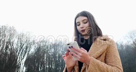 在森林附近的秋天公园年轻迷人的女人微笑着看着智能手机的肖像视频的预览图