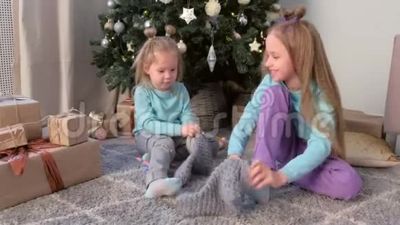 女孩们的姐妹们正在家里坐在圣诞树附近玩针织袜子视频的预览图