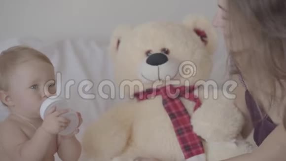 漂亮的女孩子坐在床上玩着奶瓶玩具熊就在附近视频的预览图
