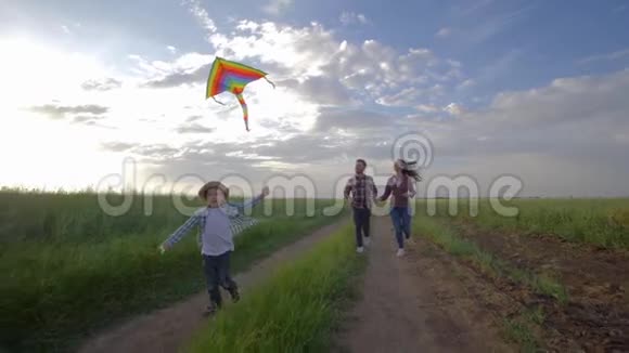 快乐的家庭周末手里拿着风筝的小男孩在乡下慢悠悠地向年轻父母跑来视频的预览图