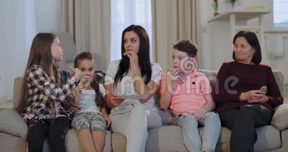 漂亮的女人带着她的三个孩子和一个奶奶在镜头前一边吃饼干一边看电影视频的预览图