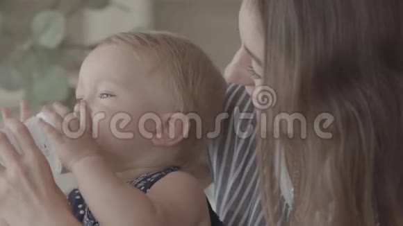 特写美丽微笑的年轻女子从厨房里的小婴儿瓶里喂她的女婴一种概念视频的预览图