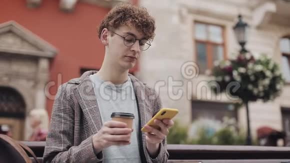 年轻的嬉皮士戴着眼镜和夹克坐在老城区的长凳上用智能手机喝着咖啡视频的预览图