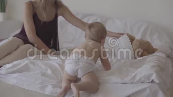 小宝宝在家在床上玩可爱的母亲坐在婴儿旁边可爱的孩子爬在床上一种概念视频的预览图