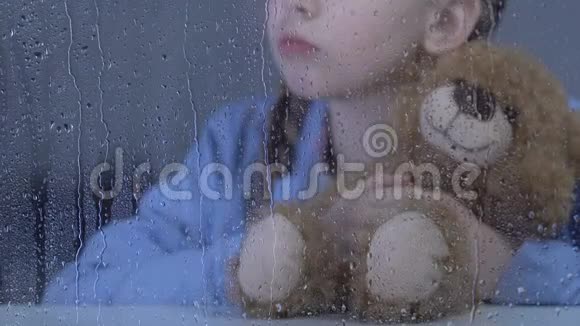 小孤独的女孩抱着玩具熊在雨窗后等待父母视频的预览图