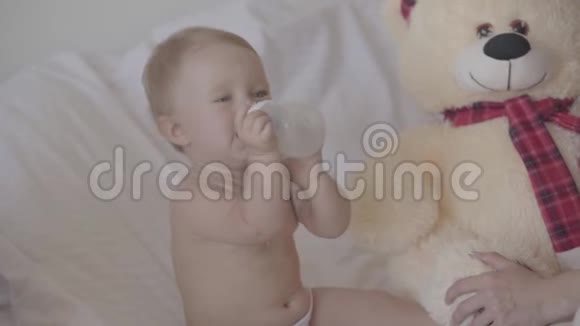 漂亮的女婴在家里坐在床上喝瓶子里的水可爱的白种人在室内视频的预览图