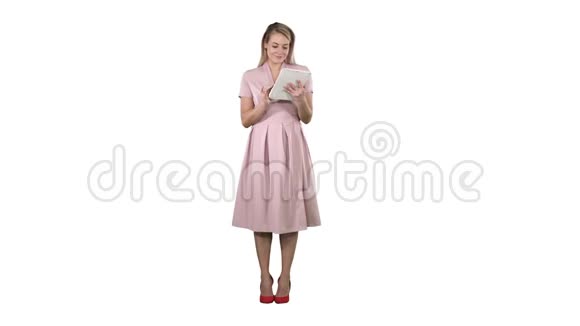 漂亮的年轻女人穿着粉红色的连衣裙拿着平板电脑在白色的背景上看着它视频的预览图