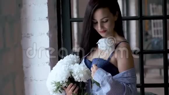 美丽的年轻女子穿着男士衬衫和胸罩站在窗边放着一束白花视频的预览图