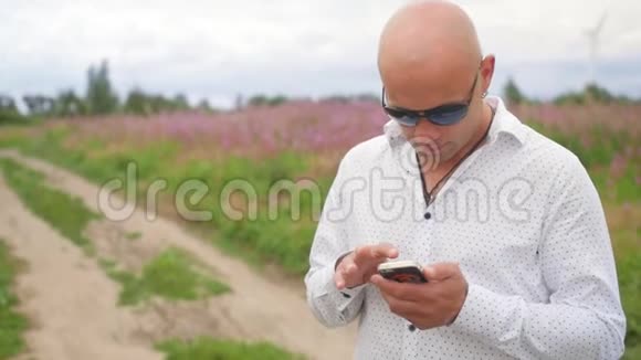 秃头男人看着电话在夏天的乡村道路和田野的背景上微笑中弹视频的预览图