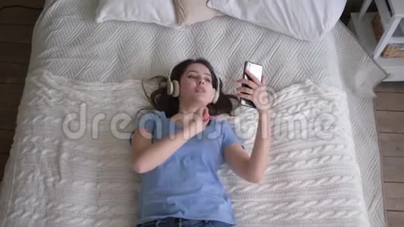 带着耳机欣赏音乐在床上放松时用手机拍照的快乐女人视频的预览图