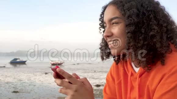 一位非洲裔美国少女年轻女性坐在海边用她的智能手机进行社交媒体或信息传递视频的预览图