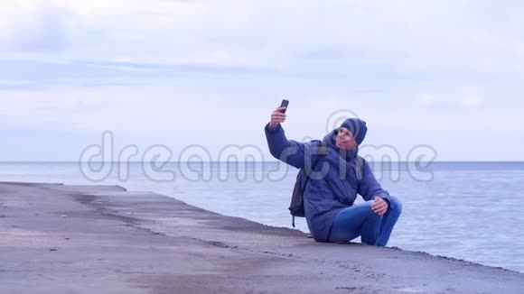 男人旅行者在智能手机上自拍冬天坐在海上的老海滨视频的预览图