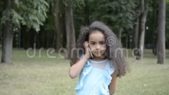 美丽的五岁非洲小女孩在一个城市公园的夏日里用手机聊天视频的预览图