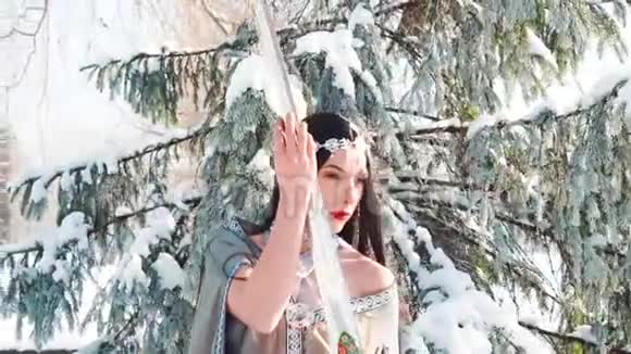 穿着灰色长斗篷穿着冬雪森林里的漂亮小姐雪皇后拿着银剑视频的预览图