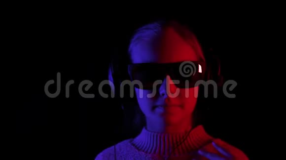 快乐女孩听音乐在耳机和黑色太阳镜在黑暗工作室与红色和蓝色照明少女视频的预览图