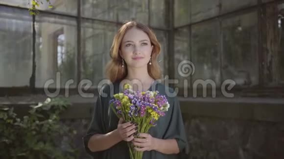 一幅美丽的年轻女子站在老楼前嗅着野花的肖像夏季休息日概念视频的预览图