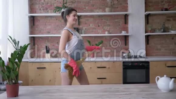 戴着手套的快乐女孩像吉他一样在扫帚上弹奏在家里打扫厨房时快乐地跳舞视频的预览图