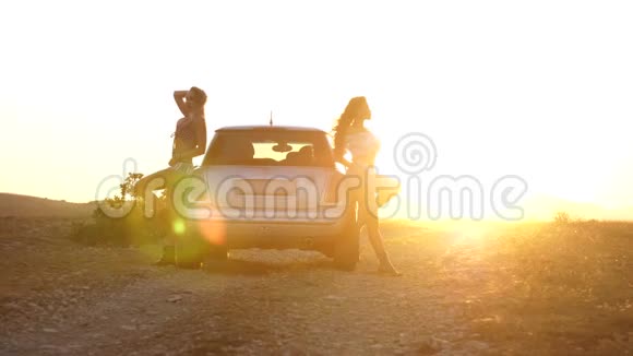 两个迷人的年轻女人站在一辆车上在光线和夕阳的照耀下在山上视频的预览图