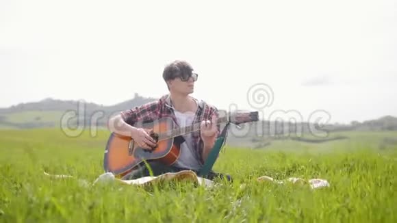 一个年轻人坐在明亮的绿色草地上的毯子上弹吉他视频的预览图