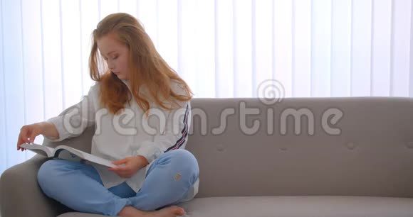 年轻可爱的白种人女孩坐在公寓里的沙发上看书的特写照片视频的预览图