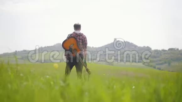 一位年轻的吉他手走在明亮的绿色草地上旅行的概念视频的预览图